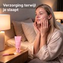 Een vrouw met een satijnen ochtendjas die collageen masker aanbrengt in een slaapkamer. Roze tube op nachtkastje.