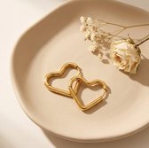 Twee gouden hartvormige oorbellen liggen op een beige schaal, naast gedroogde bloemen.