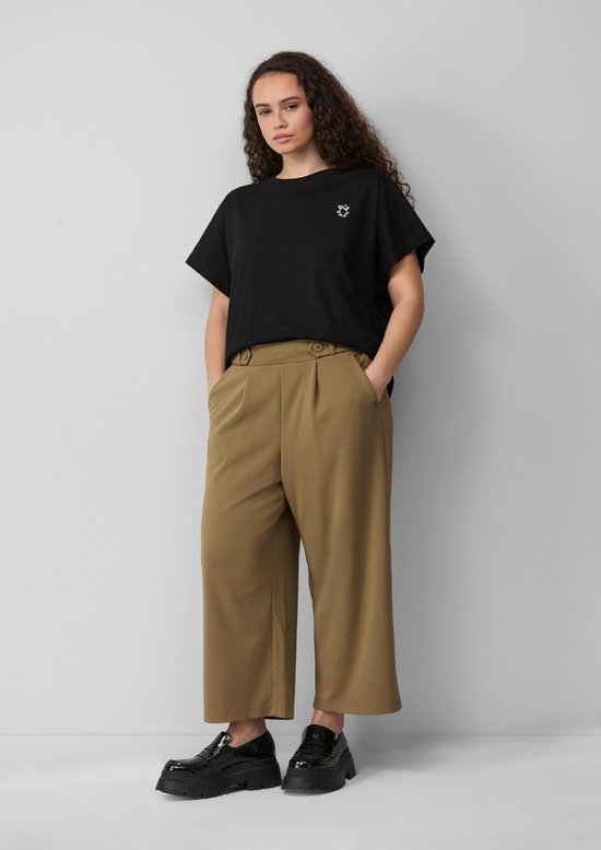 Model met lange, krullende donkere haren, draagt een zwarte top met opdruk en kaki culottebroek.