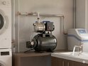 Een zwarte en zilverkleurige waterpomp met blauwe handvatten, naast een wasmachine en spoelbak in een bijkeuken.