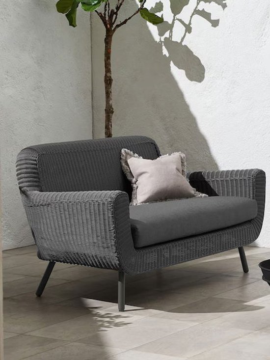 Grijze wicker bank met stoffen kussens, lichtgrijze sierkussen met franjes. Buitenomgeving met plant en schaduw.