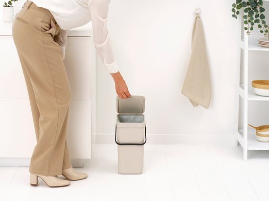 Vrouw doet afvalzak in een licht beige dubbele prullenbak met open deksels.