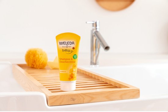Weleda baby shampoo & douchecrème in gele tube, op een houten plankje in een wastafel met badaccessoires.