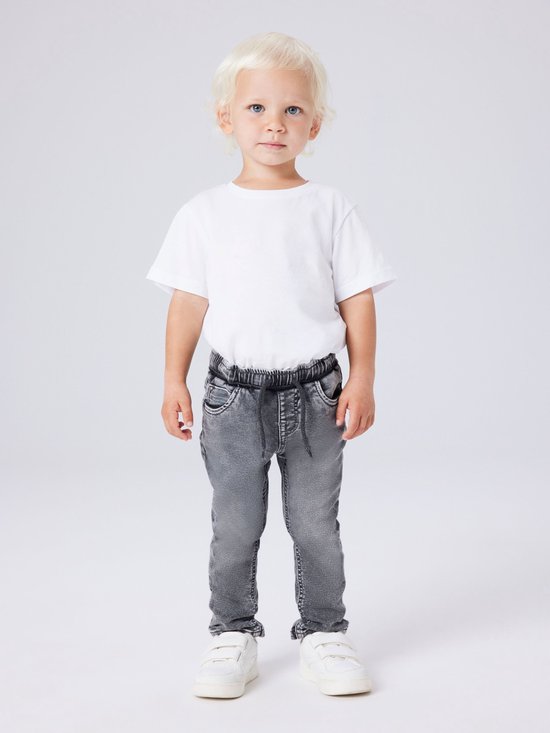Jongetje in een witte t-shirt en grijze jeans met strikceintuur en witte sneakers.