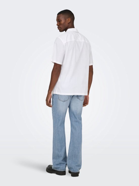 Man met witte korte mouwen blouse en lichtblauwe flared jeans, staand.