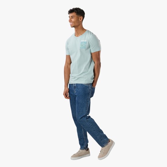 Man met blauwe jeans, lichtblauwe T-shirt met zeeschildpad-print op de borst, lachend en wegkijkend.