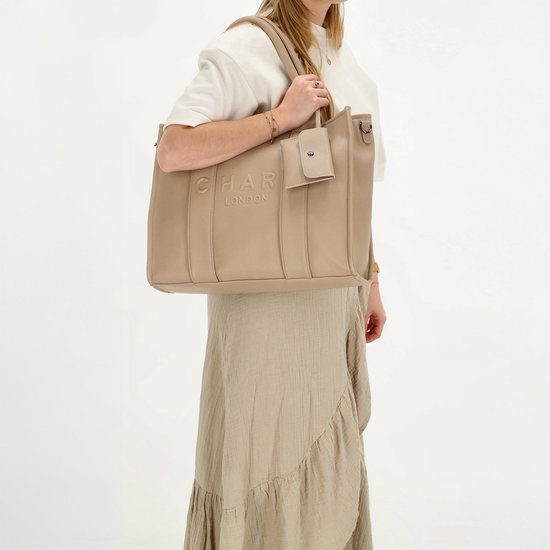 Vrouw met beige handtas met 'CHARM LONDON' logo, draagt deze crossbody met schouderband.