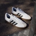 Twee witte adidas sneakers met bruine zolen en zwarte strepen op een betonnen ondergrond.