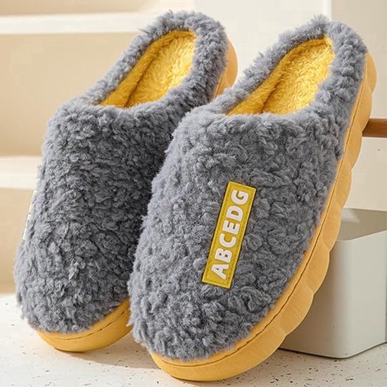 Grijze pluche pantoffels met gele binnenzool en gele badge met tekst ABCEDG.