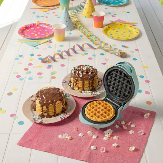 Blauw mini wafelijzer op tafel met confettie. Twee stapels wafels met chocoladesaus en toppings. Een goudbruine wafel wordt gebakken.