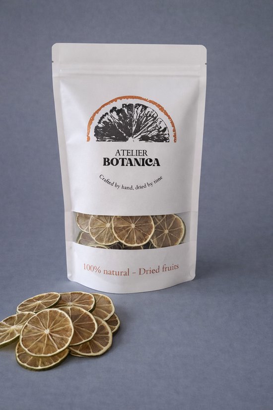 Witte zak met Atelier Botanica logo. Zichtvenster toont gedroogde limoenschijfjes. Op de voorgrond liggen limoenschijfjes.