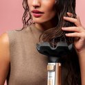 Vrouw met nat bruin haar, hand op haar haar, dichtbij een zwart diffusor-accessoire van een gouden stylingborstel.