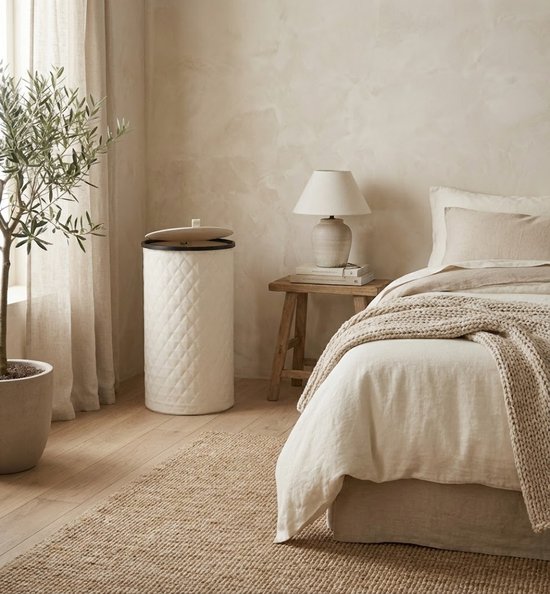 Crèmekleurige MAZAZU wasmand met ruitpatroon naast een bed in een slaapkamer.