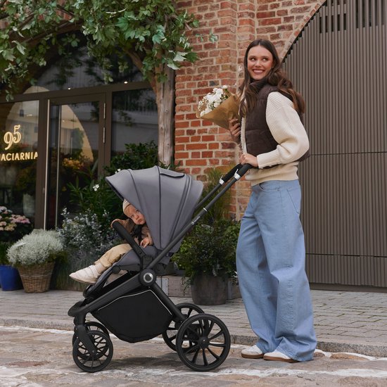 Een vrouw in lichte jeans en een vest met bruine accenten duwt een grijze kinderwagen