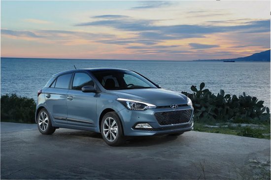 Blauwe Hyundai i20 hatchback met koplampen aan, geparkeerd aan de kust bij zonsondergang.