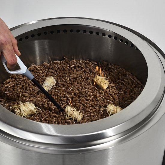 Een hand gebruikt een gasaansteker om aanmaakwokkels in een metalen vuurkorf te ontsteken, gevuld met houtpellets.