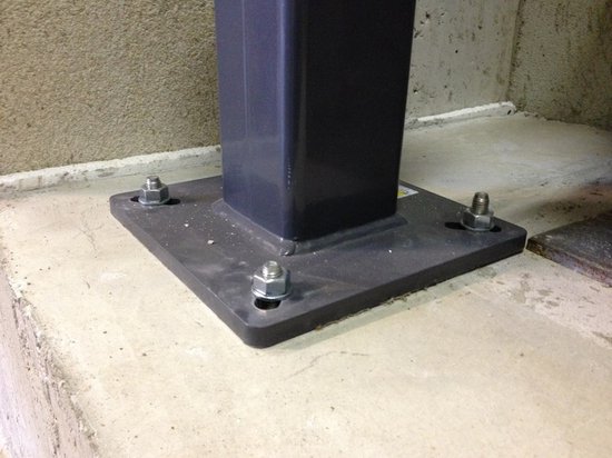 Een donkergrijs metalen frame met vier moeren en ringen bevestigd aan een betonnen vloer.