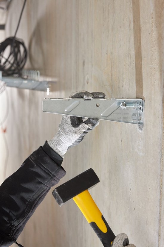 Hand met grijze werkhandschoen bevestigt een metalen anker aan een betonnen muur met een hamer.