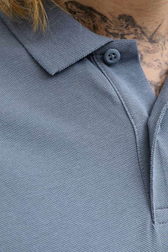 Close-up van een blauwgrijze poloshirt met een knoopsluiting en kraag. Een deel van een tatoeage is zichtbaar.
