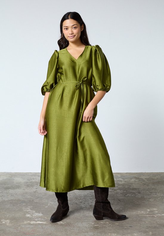 Modèle portant une robe vert olive à col en V, manches bouffantes et ceinture à nouer. Elle porte des bottes marron.