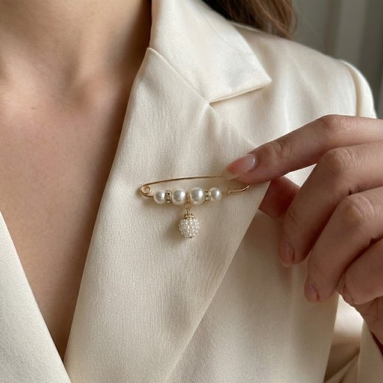 Gouden broche speld met vier witte parels en een hangend pareldetail, op een crèmekleurig jasje.