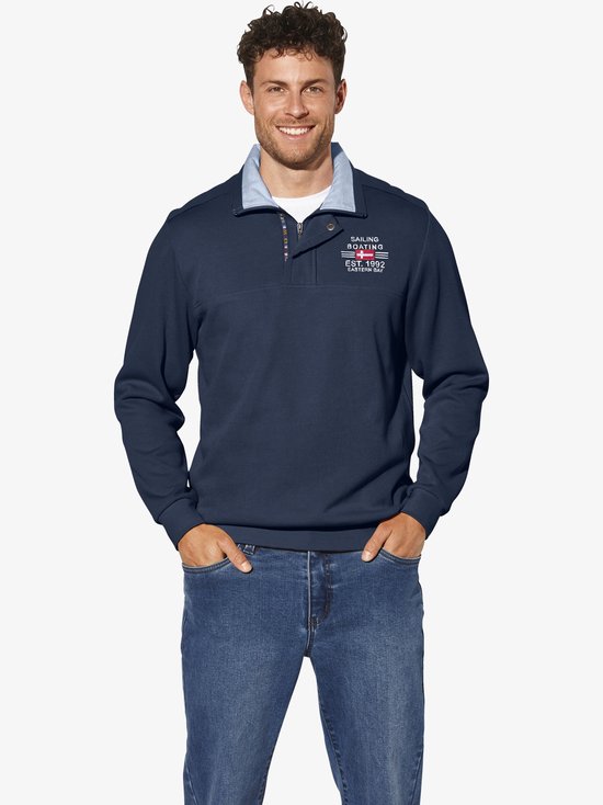 Man met spijkerbroek draagt een donkerblauwe trui met opdruk, een lichte blouse kraag en knoopjes.