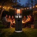 Zwarte staande terrasverwarmer met zilverkleurige details, met vlammen in de glazen buis, op een grasveld bij nacht.
