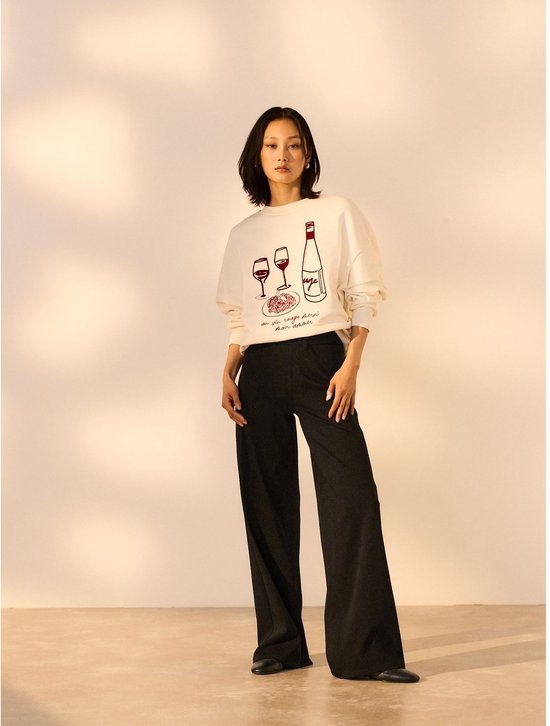 Model in een gebroken witte sweater met print van wijnglazen en een fles, gecombineerd met een zwarte broek met wijde pijpen.