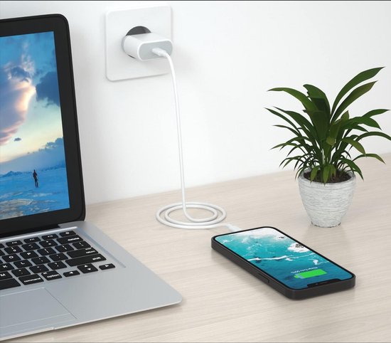 Een zilveren laptop, een witte stekker in een stopcontact en een zwarte telefoon die oplaadt. Een plant staat op de achtergrond.