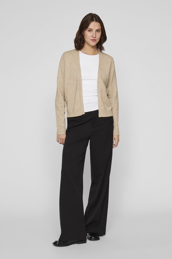 Een vrouw in een beige vest, wit T-shirt en zwarte wijde broek, staand.