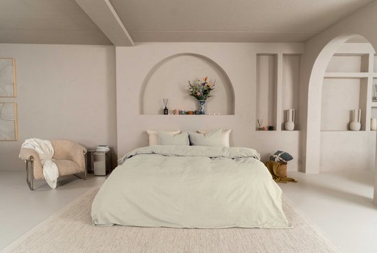 Een bed met een licht beige dekbedovertrek, lichtgrijze kussens en een fauteuil.