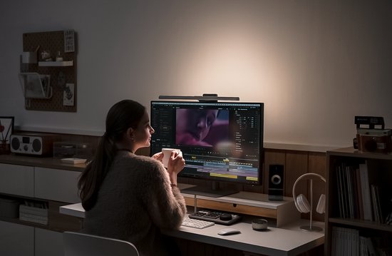 Vrouw zit aan een bureau met de BenQ ScreenBar Halo 2, kijkt naar een monitor en drinkt koffie.