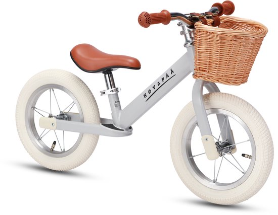 Lichtgrijze loopfiets met bruin zadel, bruine handvatten en een rotan mandje voorop.