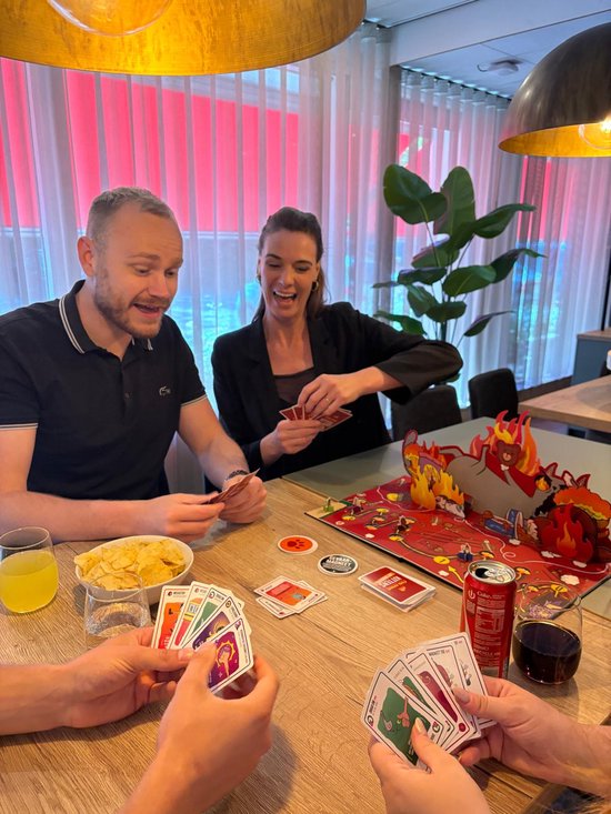 Vier spelers spelen Exploding Kittens rond een tafel met chips, drankjes en kaarten, met een pop-up speelbord op de tafel.