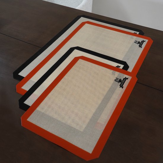 Drie siliconen bakmatten liggen op een houten tafel. Eén heeft een rode rand, één zwart en één met een crème-kleurige rand.