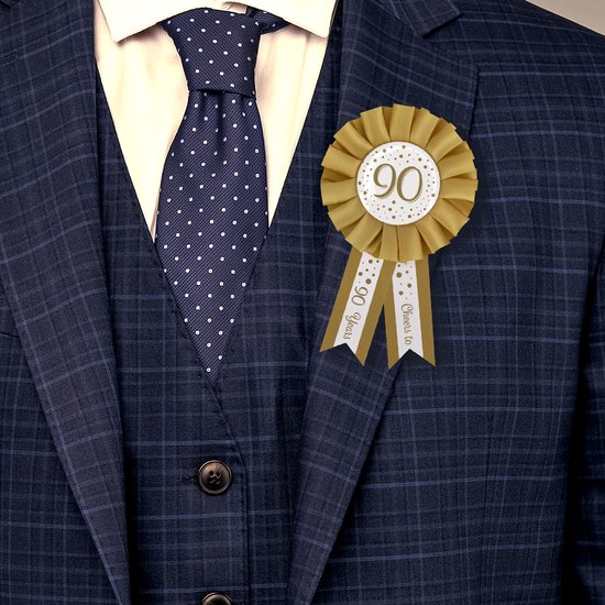Gouden rozet met "90" erop, gedragen op een pak met gestippelde das. De linten zeggen "90 Years" en "Cheers to".
