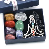 Meditatiehanger met chakra-stenen in een doos, omringd door gepolijste stenen in rood, oranje, geel, groen, blauw, indigo en paars.