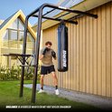 Man in sportkleding bokst tegen een zwarte bokszak, bevestigd aan een zwart metalen sportrek aan de zijkant van een houten huis.