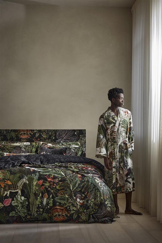 Man staat naast een bed, gekleed in een lichte badjas met jungleprint van bladeren en dieren.