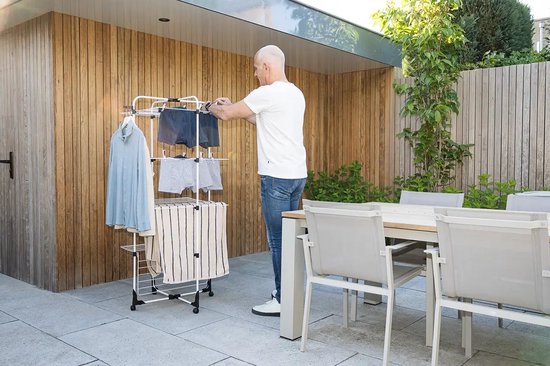 Man hangt kleding op aan een wit droogrek in een tuin met een tafel en stoelen.