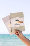 Hand houdt een bucketlist boek vast met groene kaft, met een achtergrond van de zee en strand.