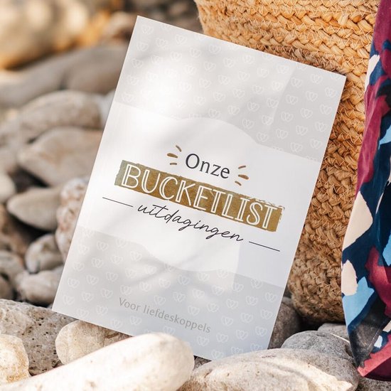 Bucketlist boek voor liefdeskoppels met hartjespatroon, gelegen naast een rieten mand en een sjaal op een stenen strand.