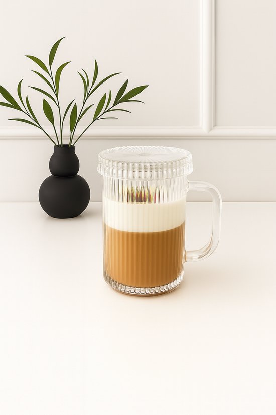 Mug en verre avec couvercle, rempli de café et de lait, à côté d'un vase noir avec une plante.