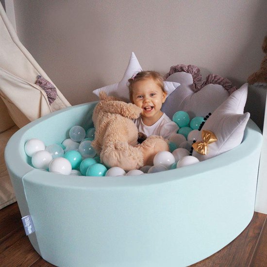 Enfant dans un parc à balles KiddyMoon vert menthe avec des balles blanches et turquoise. Coussins étoiles et ours en peluche.