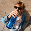 Un homme portant des lunettes de soleil et une veste en jean boit dans une gourde Kambukka bleue avec paille.