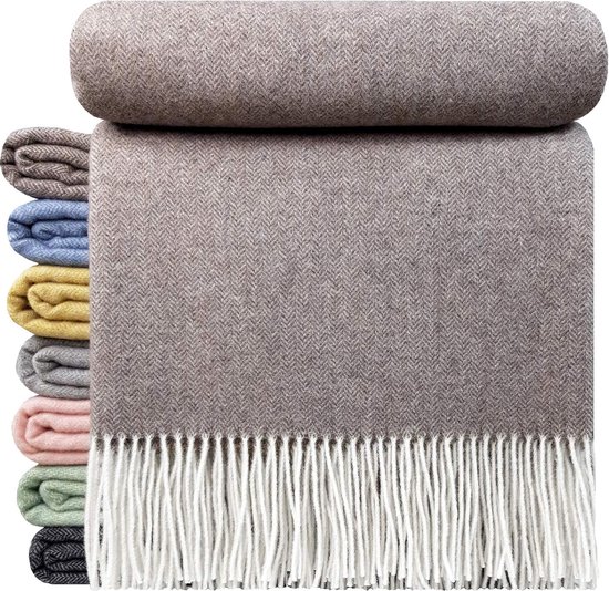 Bruine wollen plaid met visgraatmotief en witte franjes, naast een stapel kleine plaids in verschillende kleuren.
