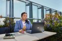 Vrouw met headset op werkt op een laptop op een balkon met uitzicht op de stad en planten.