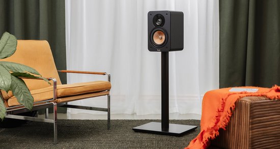 Zwarte hifi-speaker met koperkleurige woofer op een zwarte standaard, in een woonkamer met stoel.