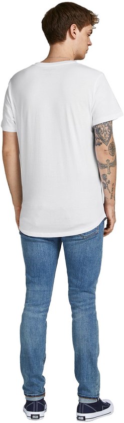 Een man met tatoeages op zijn arm draagt een witte t-shirt met korte mouwen en een ronde hals, gecombineerd met een spijkerbroek.