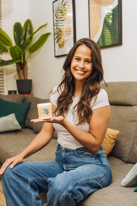 Een lachende vrouw met bruin haar, zittend op een bank. Ze draagt een wit t-shirt en spijkerbroek en houdt een potje Blije Voeten eelt- en klovencrème vast.